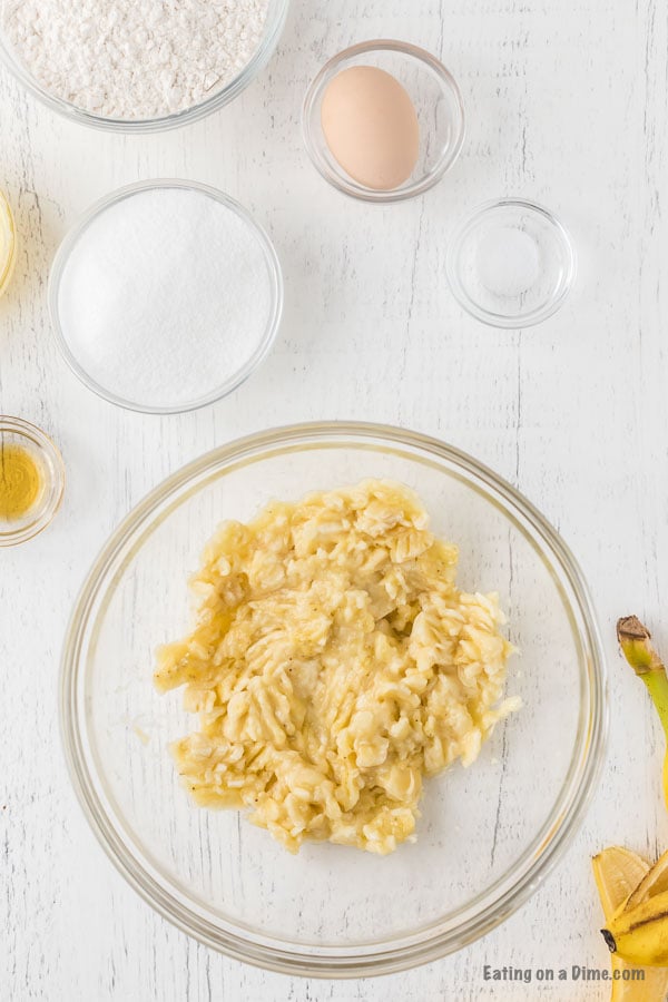 Mashing bananas in a bowl with bowls of flour, sugar, egg, and salt on the side