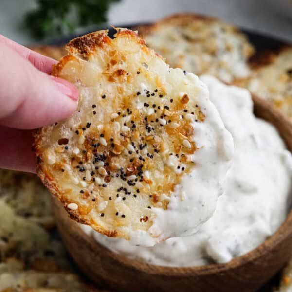 A hand holds a round Cottage Cheese Chip topped with seeds and cheese, dipping it into a bowl of creamy white dip. Other crisps are visible in the background.